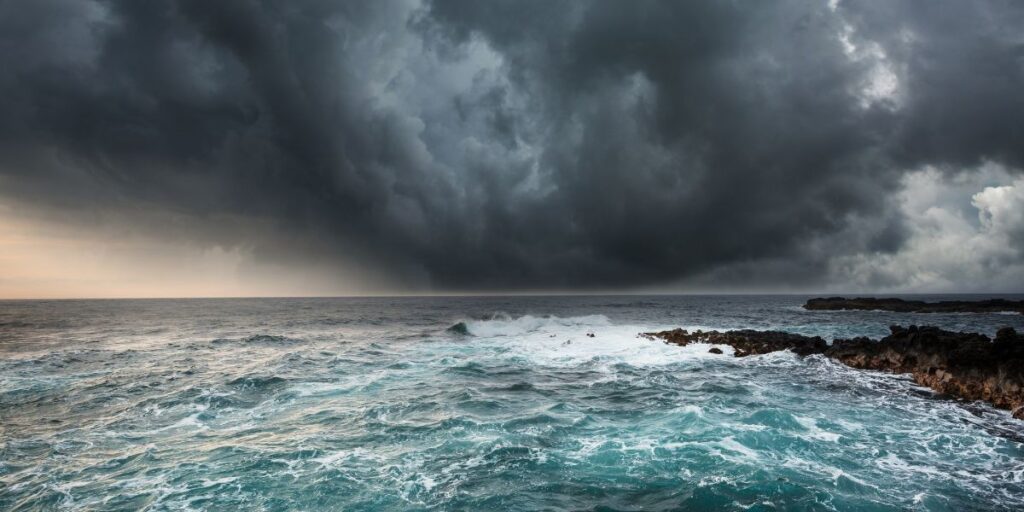 The Mediterranean Sea reveals its wild side as tumultuous waves crash against rocky shores under a brooding and storm-laden sky, a stark reminder of nature's unpredictable temperament. The contrast between the dark clouds and the sun's faint glow near the horizon adds drama to the scene.