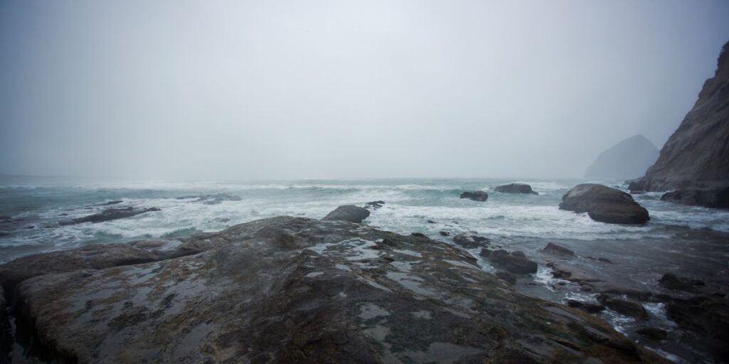 The vast expanse of the Pacific Ocean is partially shrouded by a misty haze, with waves lapping over weathered rocks near the shore. The overcast sky and the soft, diffused light give the seascape a tranquil, yet mysterious ambiance.