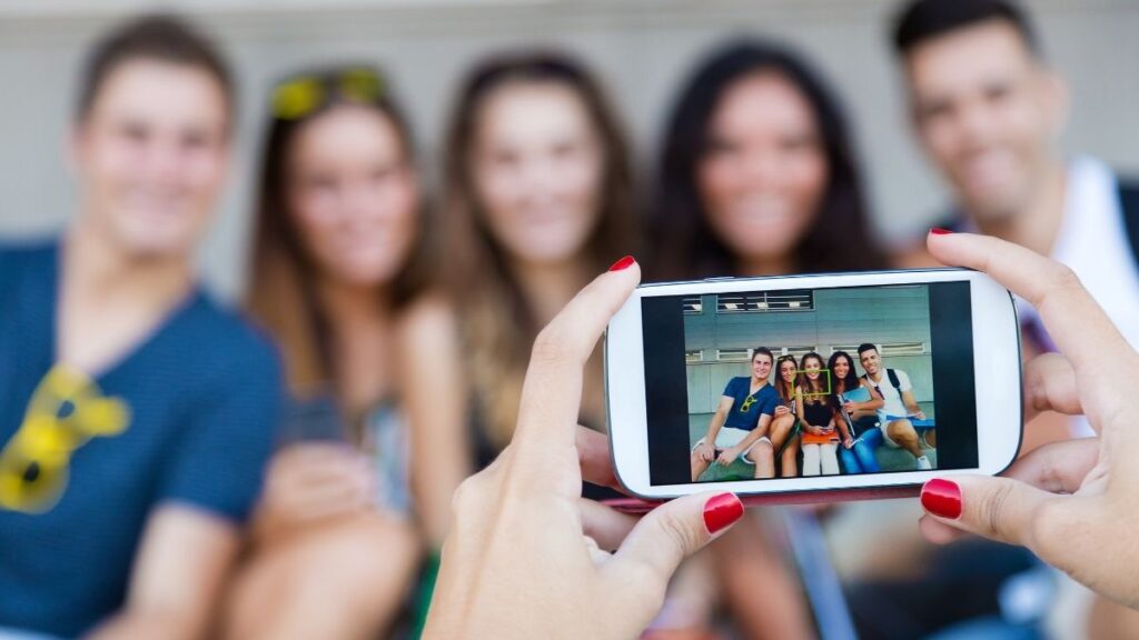 A person with red nail polish holds a smartphone, capturing a clear photo of five smiling friends posing together, while the actual group in the background is blurred. The image on the phone screen is sharp and well-framed, contrasting with the out-of-focus scene beyond. The setting appears to be outdoors or in a public space, emphasizing a fun, social moment.