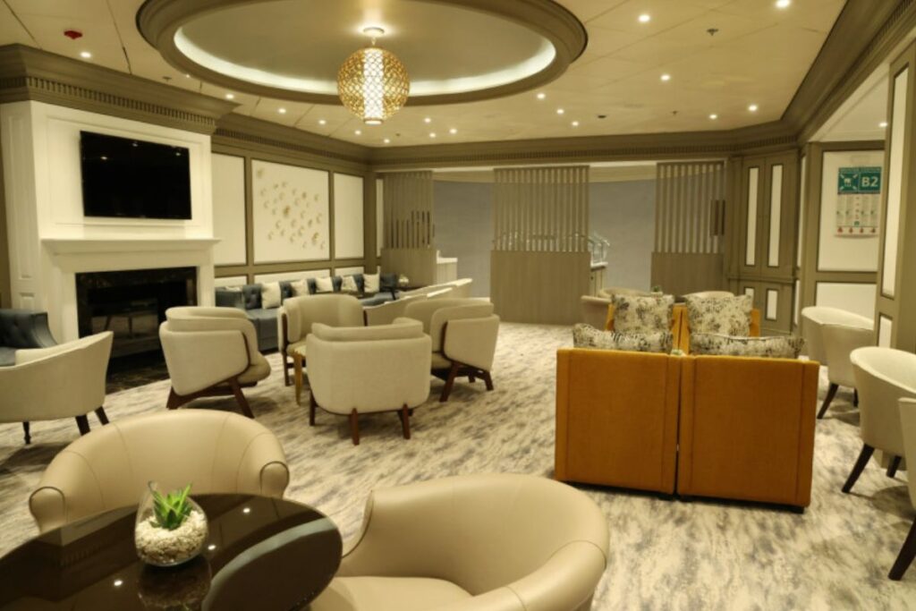 This image showcases The Retreat Lounge on the Celebrity Infinity, a sophisticated space designed for relaxation. The room features elegant seating arrangements with plush armchairs, couches, and a cozy fireplace. Soft lighting and neutral tones create a calming and luxurious atmosphere. The modern decor, including a stylish central light fixture and minimalist artwork, adds to the lounge's inviting and upscale vibe.