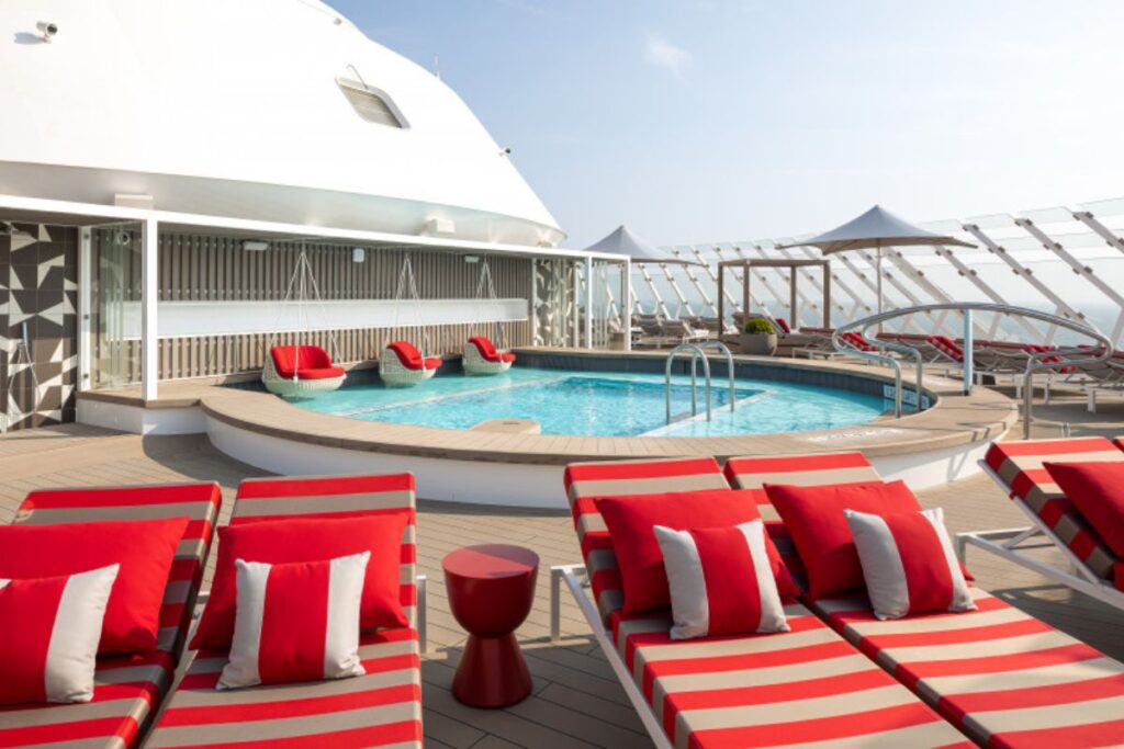 This image showcases The Retreat on Celebrity Beyond, a luxurious private lounge area featuring a pool surrounded by vibrant red and white striped lounge chairs. Cozy red cushioned hanging chairs add to the chic, relaxing atmosphere. The design is sleek and modern, with open, airy spaces and soft sunlight bathing the pool deck, creating a tranquil and exclusive ambiance for relaxation and leisure.