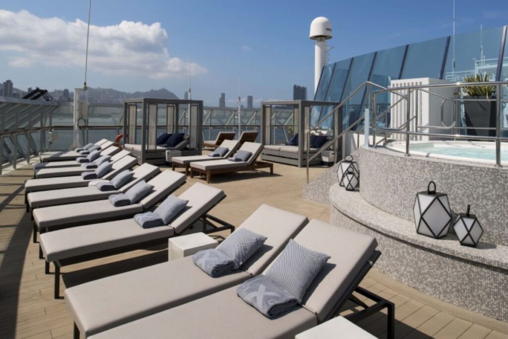This image shows The Retreat on the Celebrity Millennium, an exclusive lounge area featuring luxurious sun loungers neatly arranged around a serene deck. The space is adorned with comfortable pillows and towels, offering a chic and relaxed atmosphere. A Jacuzzi is visible in the background, along with private cabanas for extra comfort. The clear skies and distant city skyline create a peaceful yet elegant setting for guests to unwind.