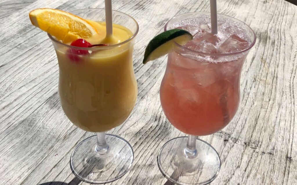 Two tropical drinks on a wooden table, one creamy yellow with an orange slice and cherry, and the other pink over ice with a lime wedge.