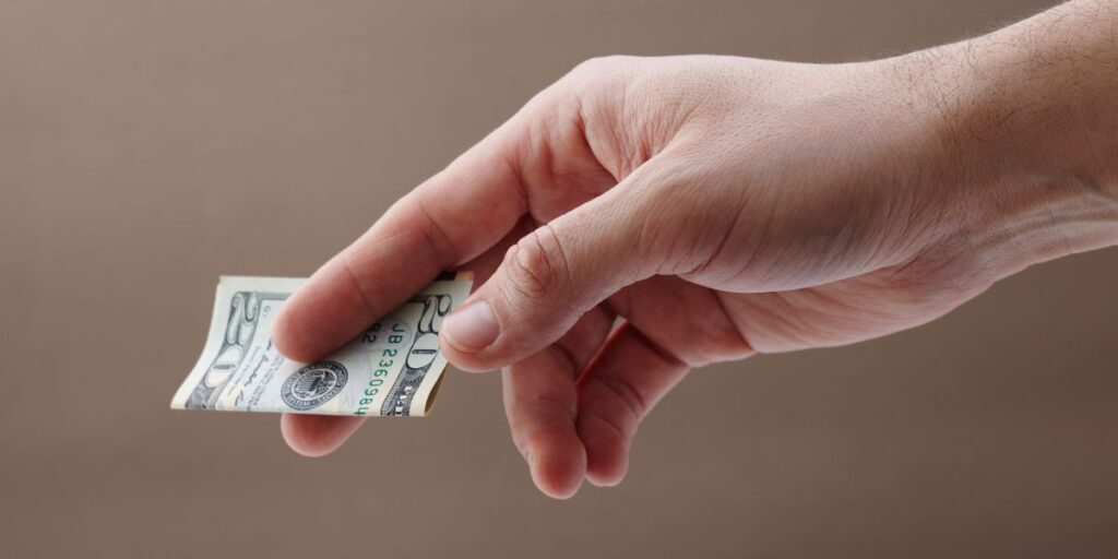 A close-up image of a hand holding a small folded stack of U.S. dollar bills, suggesting a cash transaction or a tip. The background is a plain, neutral color.