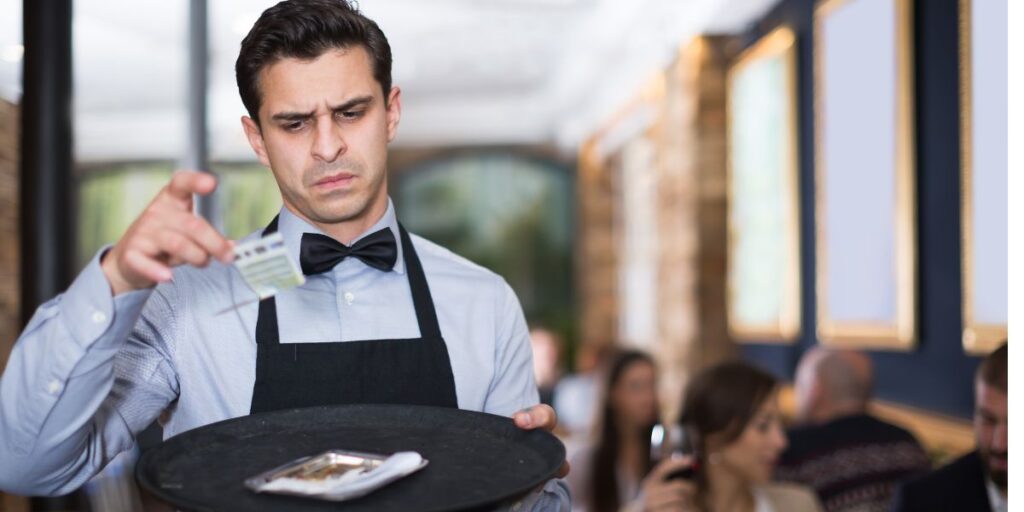 A waiter in a black apron and bow tie looks displeased as he examines a small tip on a tray in a busy restaurant. The blurred background shows patrons dining, with one holding a glass of wine.