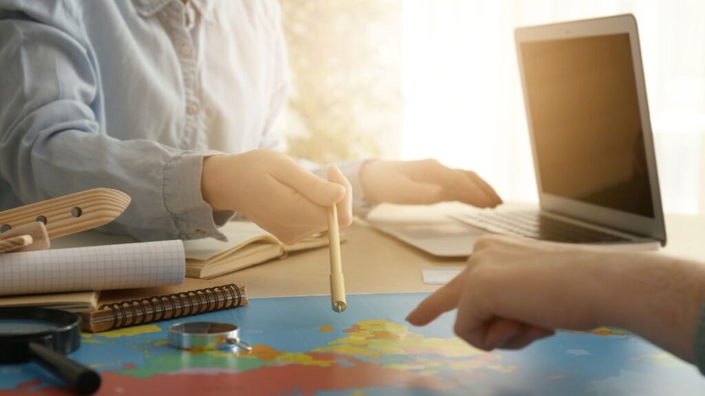 This image features a close-up of two people planning a trip, with one pointing at a world map and the other holding a pen. A laptop, notebook, and magnifying glass are visible on the table, emphasizing a collaborative travel or research setting.