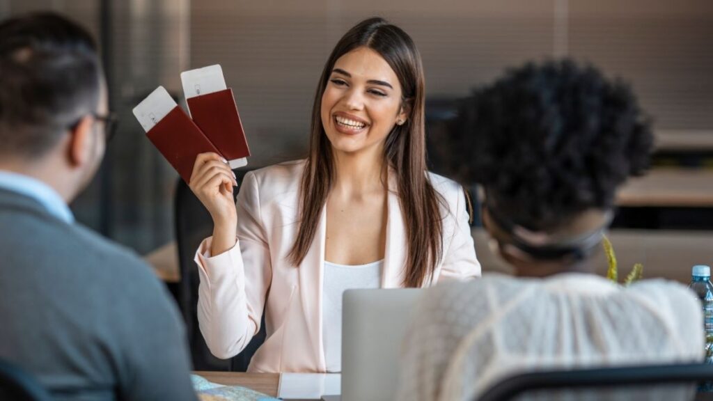 A smiling travel agent wearing a light pink blazer holds up two passports with boarding passes while sitting at a desk. Two clients, seen from behind, are seated across from her, suggesting a travel planning session.