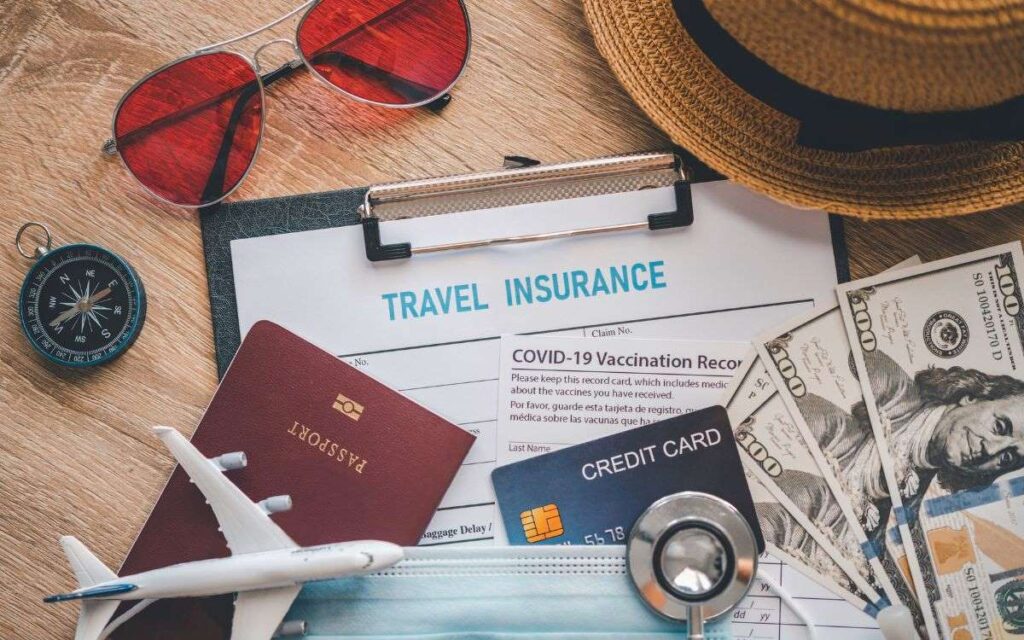 Essential travel items arranged on a wooden surface, including a passport, travel insurance documents, COVID-19 vaccination record, credit card, cash, sunglasses, a straw hat, a compass, a model airplane, and a face mask, symbolizing preparedness for travel during the pandemic.