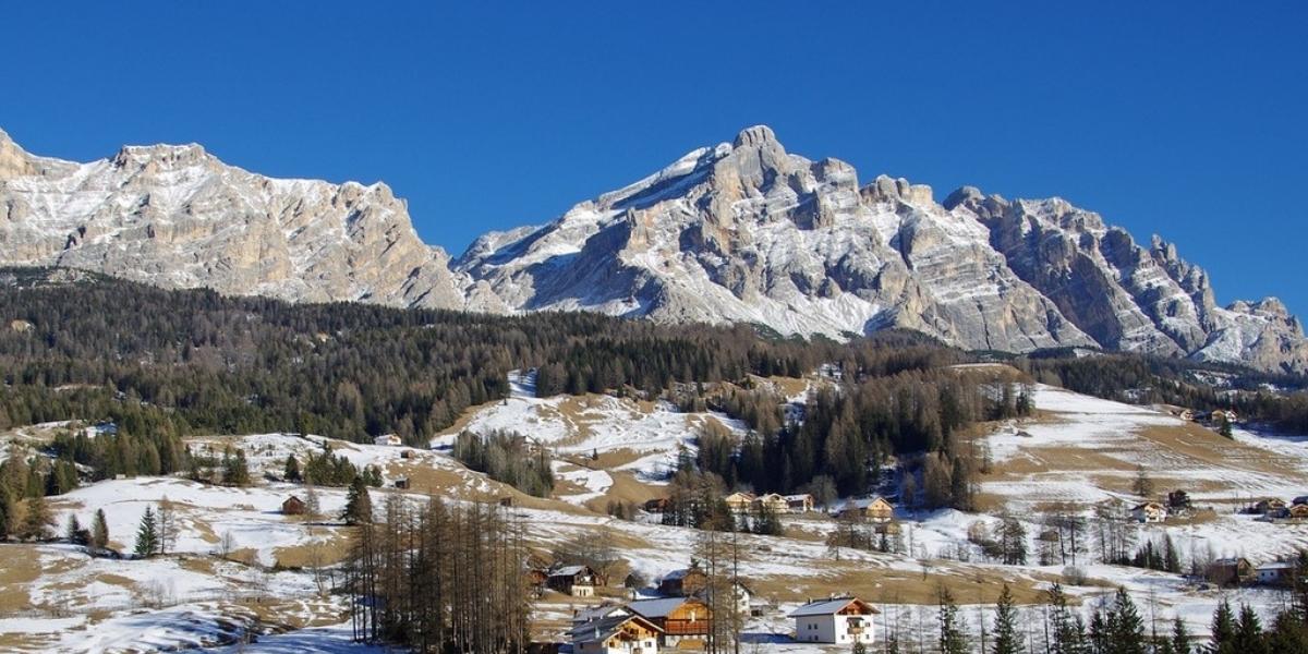 Snow-dusted alpine village nestled in the foothills of the dramatic Dolomite Mountains in northern Italy, with scattered cabins, pine trees, and rugged peaks under a clear blue sky. The scene blends early winter snow with golden patches of dry grass across gently sloping terrain.