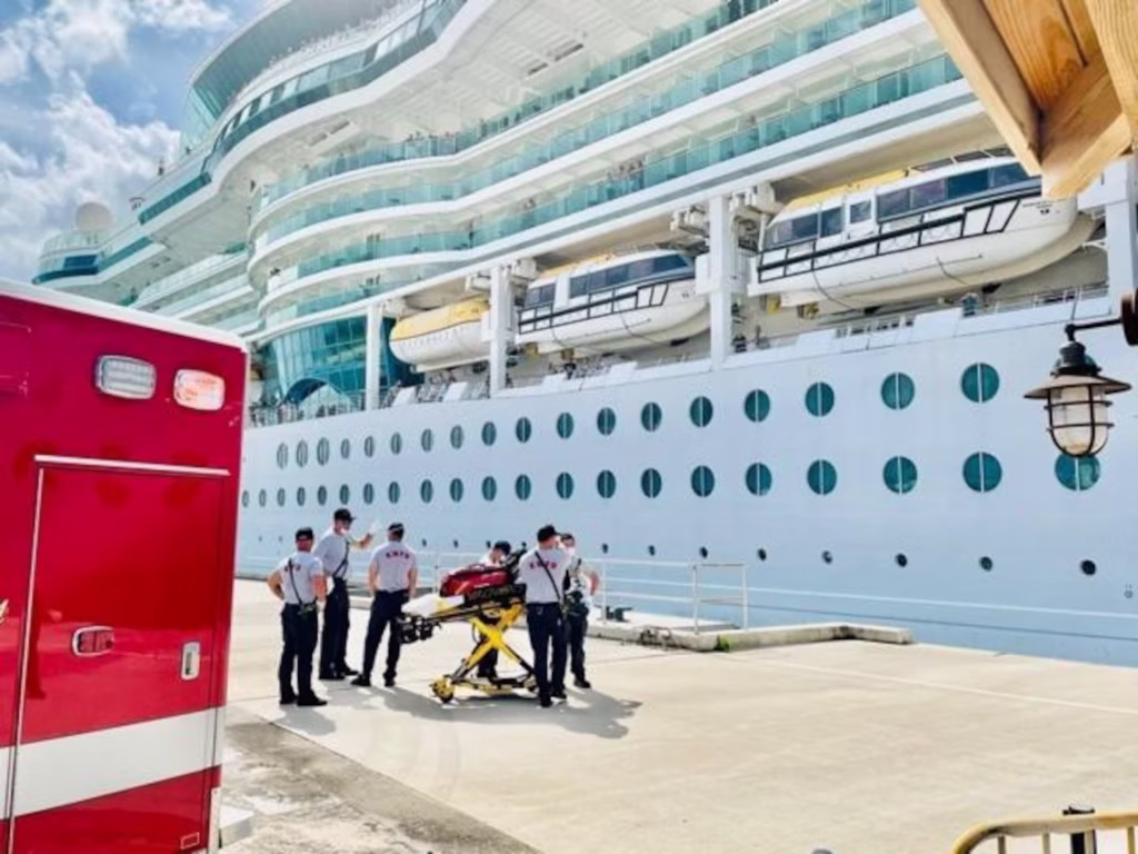Emergency responders with a stretcher beside a docked cruise ship, with an ambulance nearby at the port.