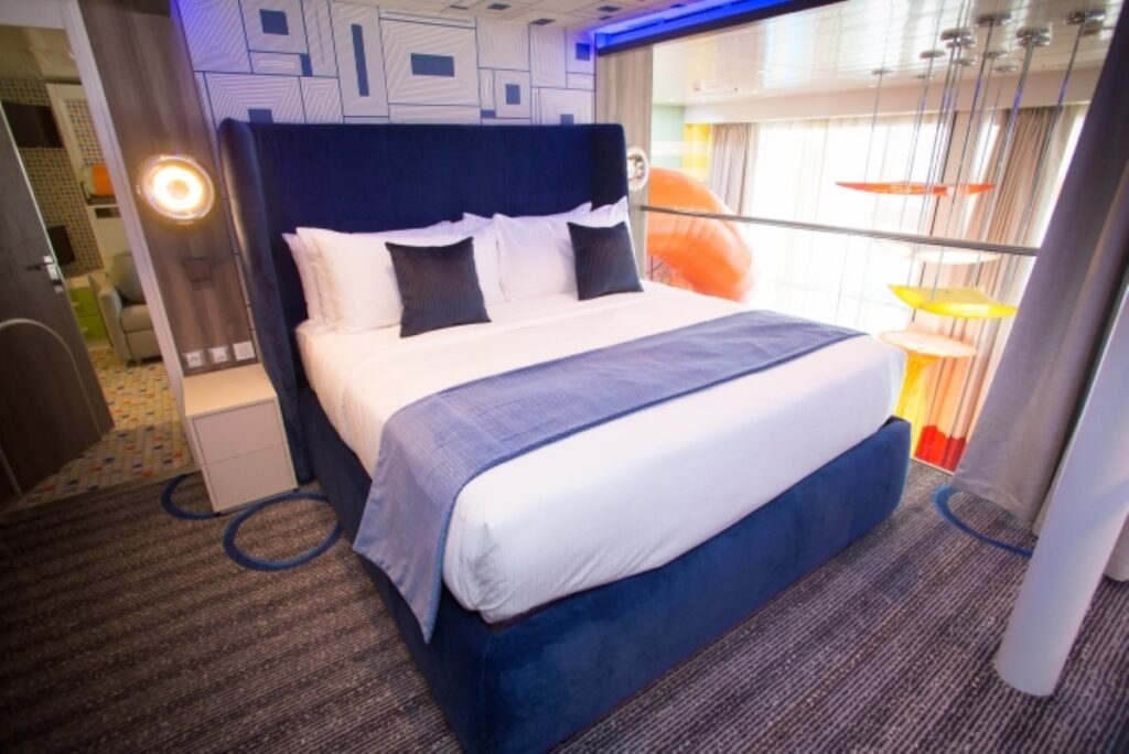 The Ultimate Family Suite on Royal Caribbean's Symphony of the Seas features a spacious, modern bedroom with a large blue headboard, plush bedding, and a view of the colorful balcony area with a swing and table tennis.