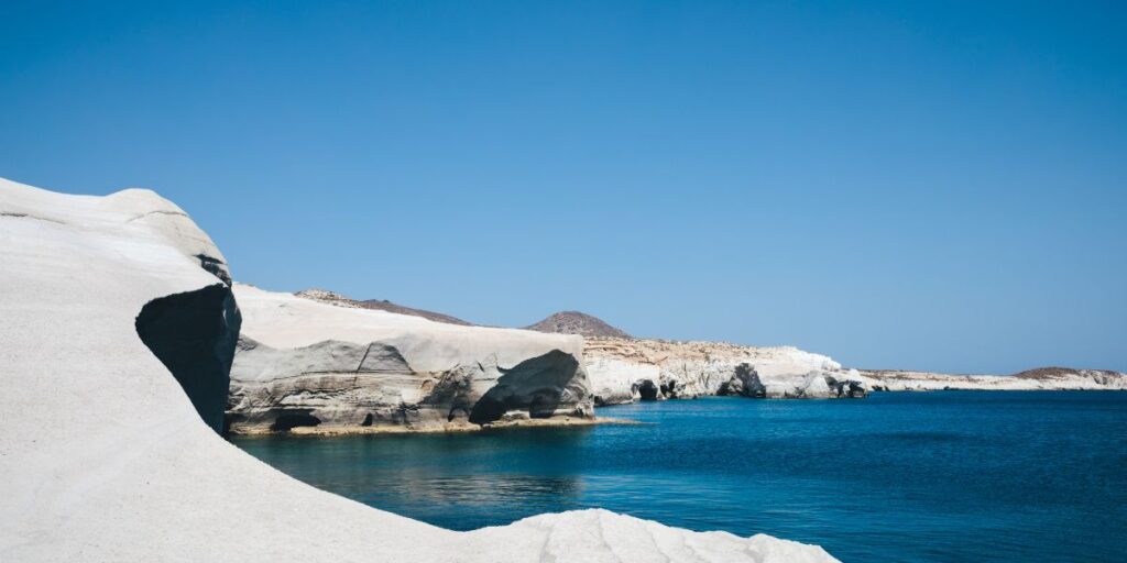 White volcanic rock cliffs curve around a tranquil turquoise bay under a cloudless blue sky. The unique smooth formations and deep blue water suggest a serene coastal landscape, likely on a Greek island like Milos.