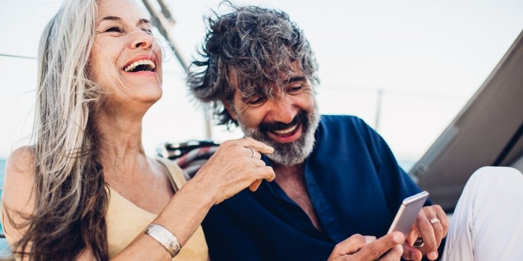 Smiling older couple relaxing on a boat, laughing together while looking at a smartphone. The woman is wearing a sleeveless top and silver bracelet, and the man is in a navy shirt, creating a carefree, vacation vibe.