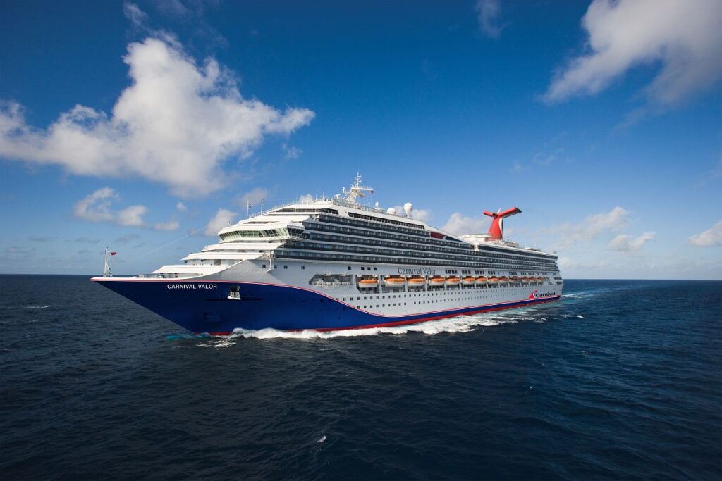 Carnival Valor cruise ship sailing on the ocean, showcasing its distinctive blue and white exterior with a red funnel, against a bright blue sky with fluffy clouds.