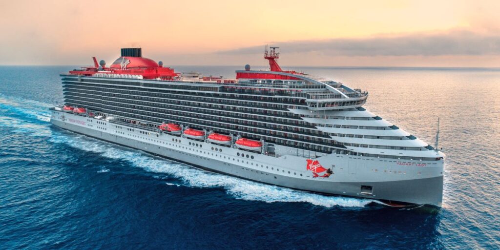 Virgin Voyages' Valiant Lady cruise ship sails across calm ocean waters at sunset. The sleek grey-and-red vessel features modern balconies, bold red lifeboats, and the signature Virgin mermaid logo on its hull.