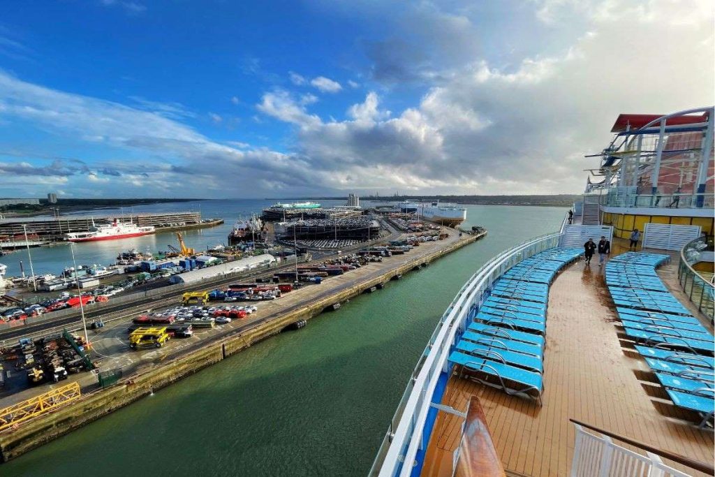 View from the deck of a Carnival cruise ship, overlooking a bustling port with rows of parked cars and industrial activity, beneath a dynamic sky with scattered clouds.