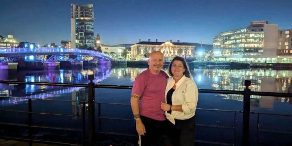 A couple poses by a waterfront railing at night, with vibrant city lights reflecting off the calm river behind them. A lit pedestrian bridge and modern buildings, including one labeled “Royal Mail,” frame the scenic urban backdrop.