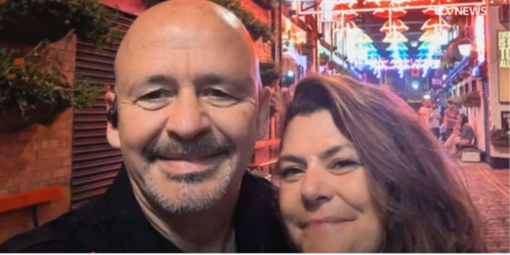 A smiling man and woman pose for a selfie in a lively, narrow alleyway lit with colorful neon umbrella lights overhead. The cobblestone street and warm atmosphere suggest a festive nightlife setting, with people casually gathered in the background.