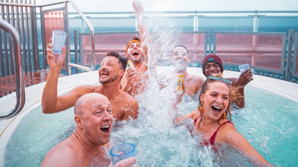 A group of six people is having a lively time in a jacuzzi on a Virgin Voyages cruise. They are splashing water, laughing, and holding drinks while enjoying the moment. The background shows an ocean view with glass railings, giving a luxurious and carefree vacation vibe.