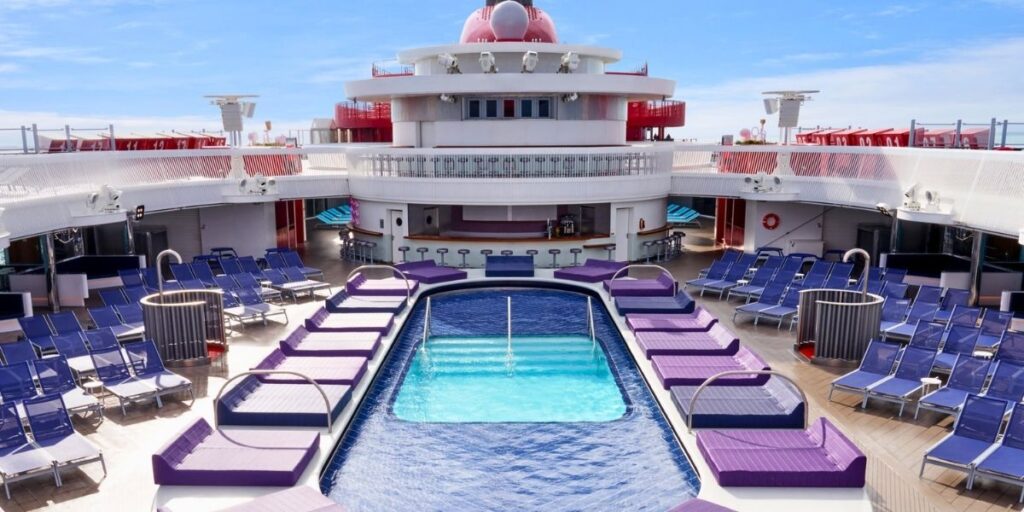 This image shows the lido deck of a Virgin Voyages cruise ship, centered around a sleek rectangular pool. Surrounding the pool are modern purple cushioned loungers and rows of blue reclining sunbeds, with a bar area and shaded seating in the background, all under a bright blue sky.
