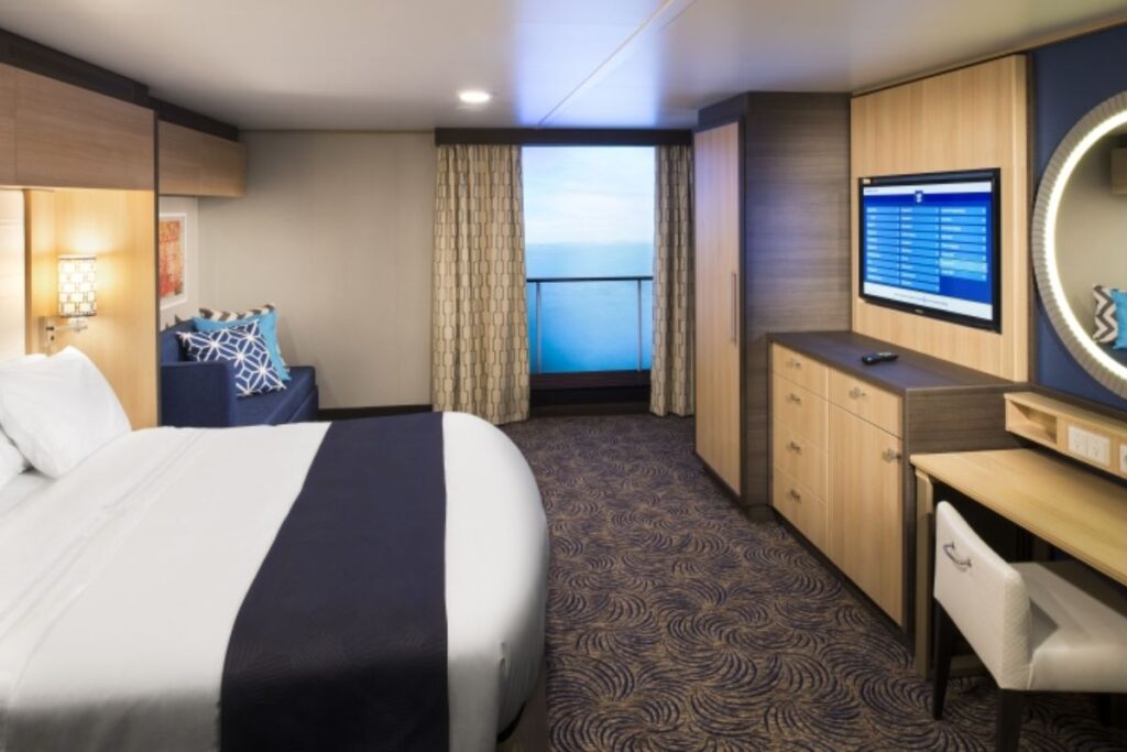 This image shows a stateroom on Anthem of the Seas featuring a Virtual Balcony. The room is designed with modern, neutral tones and includes a comfortable bed, a sitting area with accent pillows, and a flat-screen TV mounted on the wall. The standout feature is the Virtual Balcony, which simulates an ocean view using a large screen framed by curtains, offering an immersive experience even in interior cabins. The room’s sleek furnishings create a cozy yet contemporary space for relaxation.