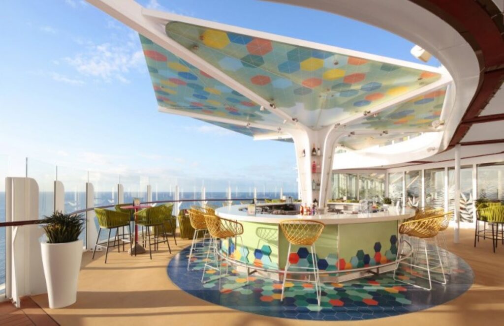 The Vue Bar on Royal Caribbean’s Wonder of the Seas, bathed in natural light, boasting a colorful, modern design with a mosaic-tiled bar and whimsical overhead canopy. The outdoor bar offers a panoramic ocean view, accented with stylish chairs and a relaxed, contemporary ambiance.