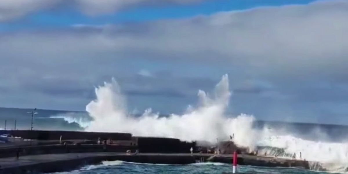 A towering wave crashes over a concrete pier in Tenerife, sending a huge spray of seawater into the air as several people stand nearby watching the surf under a cloudy sky.v