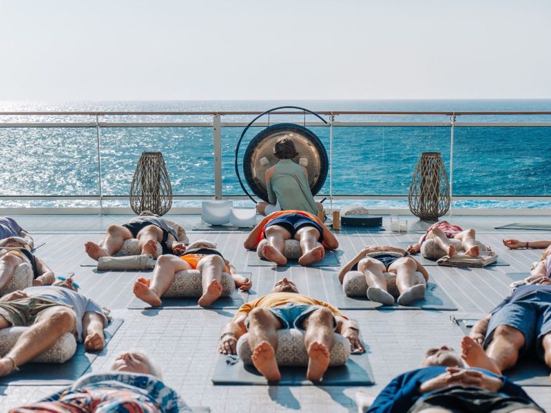 Cruise guests participate in a relaxing outdoor wellness session on deck as an instructor uses a gong with the ocean in the background.