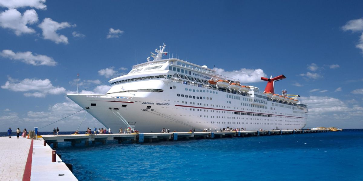 Carnival cruise ship docked at a port with clear blue skies and vibrant turquoise waters, with passengers strolling on the pier.