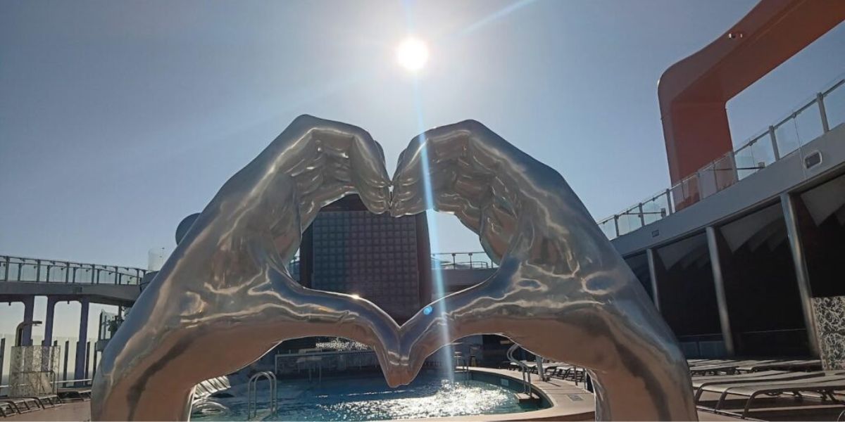 A modern sculpture of two hands forming a heart shape with a sun shining brightly above. The sculpture is positioned on the deck of the Celebrity Apex cruise ship, creating a picturesque and artistic scene.