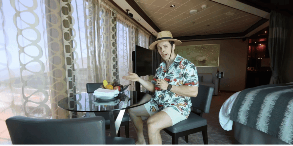 A man in a straw hat and tropical shirt sits at a small dining table inside a cruise ship suite, pointing toward a fruit bowl with bananas, apples, grapes, and oranges. Sunlight filters through sheer, patterned curtains behind him, adding to the relaxed vacation vibe of the room.