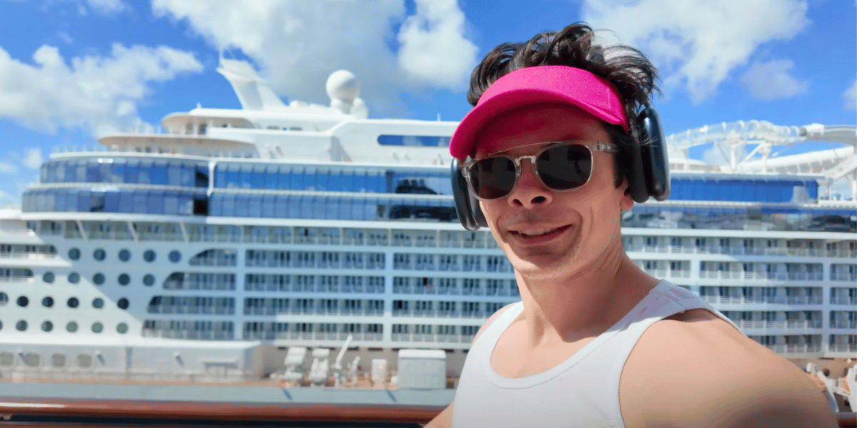 Will Tennyson wearing a bright pink visor, large headphones, sunglasses, and a white tank top takes a selfie in front of a massive white cruise ship under a sunny blue sky. The ship’s upper decks and glass balconies are clearly visible in the background.