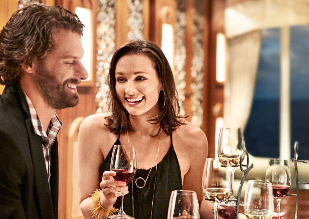 The image features a couple enjoying a wine tasting experience on a Princess Cruise. They are both holding glasses of red wine, engaging in cheerful conversation with a sense of intimacy and enjoyment. The setting is luxurious, likely a dining or lounge area on the ship, with an elegant decor and a window that suggests an ocean view, contributing to a romantic and sophisticated ambiance.