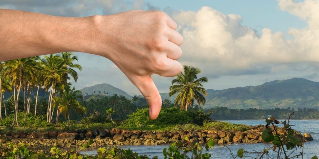 A large hand in the foreground gives a thumbs-down gesture over a scenic tropical island landscape with palm trees, rocky shoreline, and distant mountains under a partly cloudy sky. The gesture contrasts humorously with the otherwise picturesque setting.