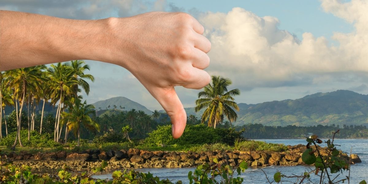 A large hand in the foreground gives a thumbs-down gesture over a scenic tropical island landscape with palm trees, rocky shoreline, and distant mountains under a partly cloudy sky. The gesture contrasts humorously with the otherwise picturesque setting.