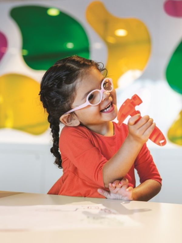 A cheerful young girl with glasses, wearing a red shirt, joyfully playing with a toy in the colorful Kid's Club area aboard the Anthem of the Seas cruise ship.