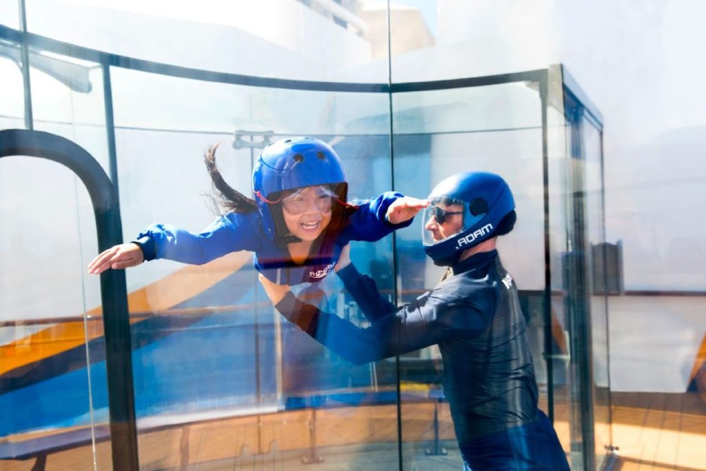 A young girl in a blue flight suit and helmet is smiling with excitement during a simulated skydiving experience in the RipCord by iFly attraction on a Royal Caribbean cruise ship, with an instructor guiding her.