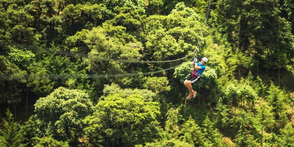 An exhilarating scene of a person zip-lining over a dense rainforest canopy, dressed in a safety harness and helmet, with a vast expanse of vibrant green trees below them, showcasing an adventurous outdoor activity.