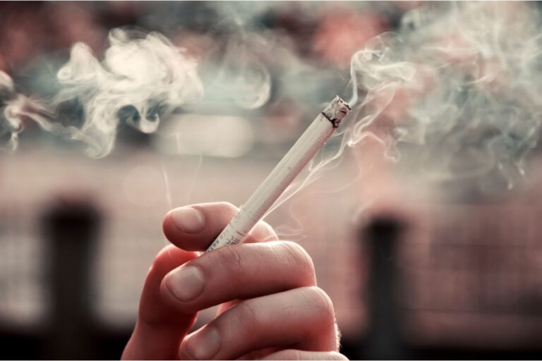 A close-up image of a person's hand holding a lit cigarette, with smoke curling upwards. The focus on the cigarette and the smoke against a blurred background emphasizes the act of smoking.