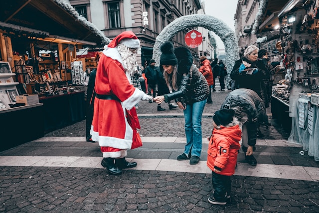 Santa Claus in a red suit hands out treats to a family at a festive Christmas market in a European town on a snowy city street.