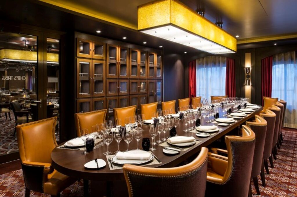 Elegant dining setup at the Chef's Table on Quantum of the Seas, featuring a long table with leather chairs, sophisticated place settings, and a warm, intimate lighting overhead. A backdrop of dark wood shelves filled with wine glasses adds to the exclusive atmosphere of the venue.