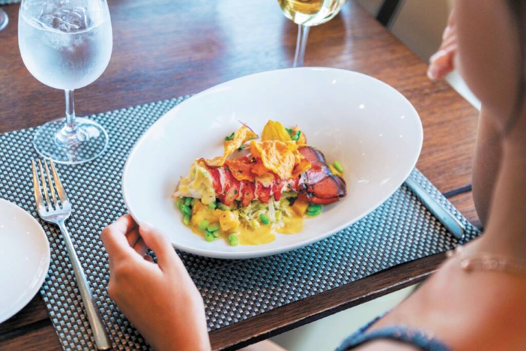 A guest about to enjoy a gourmet meal at the Coastal Kitchen on Anthem of the Seas, featuring a beautifully plated lobster dish accompanied by a chilled glass of water and a glass of white wine.