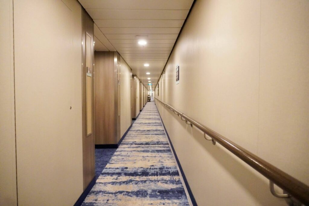 Long cruise ship hallway with cabin doors, overhead lights, a handrail along the wall, and a blue patterned carpet stretching into the distance.