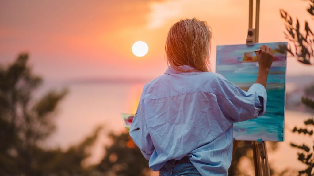 A lady painting a picture on an easel with the sun setting in the background over the sea