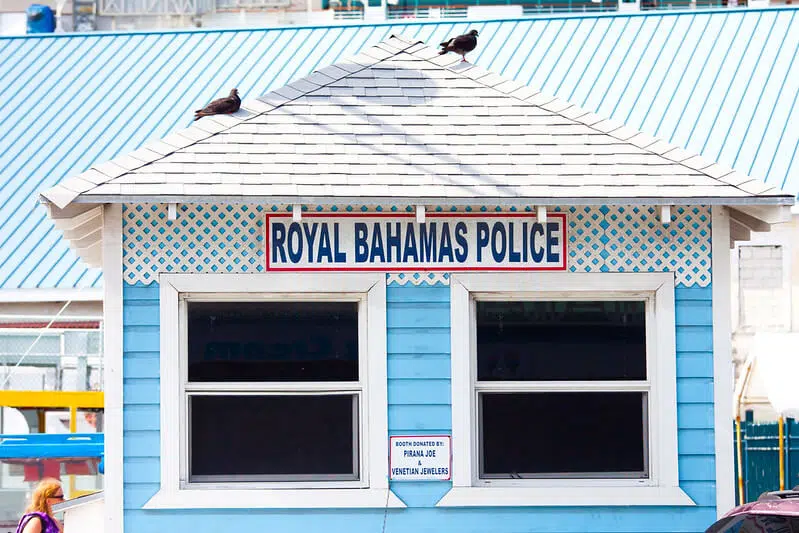 A small light blue police booth labeled “Royal Bahamas Police” with two windows and a white shingled roof, topped with pigeons. The booth sits in a tropical port area, featuring a colorful, coastal architectural style.