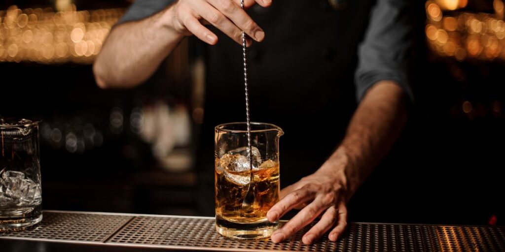 A bartender uses a long stirring spoon to mix a cocktail in a glass beaker filled with ice cubes, set against the backdrop of a dimly lit bar. The bartender's hands and the beaker are in focus, while the background features a warm, bokeh effect from the bar lights.