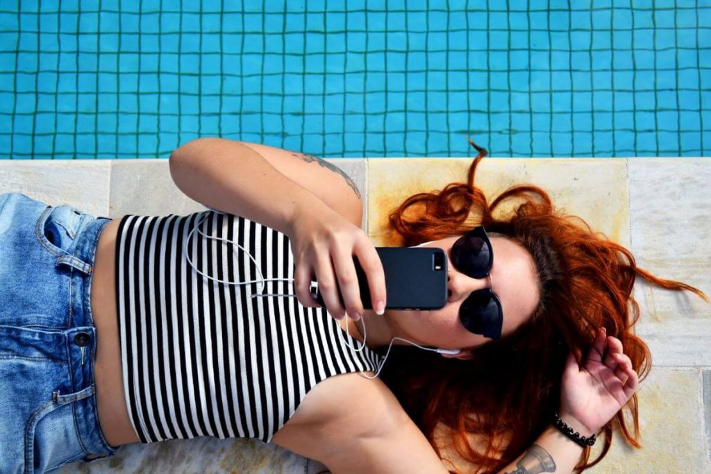 A woman in a striped tank top and denim shorts relaxing by the poolside, taking a selfie with a smartphone while wearing sunglasses and earphones.