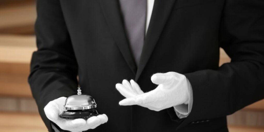 A cruise butler dressed in a black suit with white gloves holds a silver service bell in one hand while gesturing with the other. The background is softly blurred, highlighting the formality and attention to service detail.