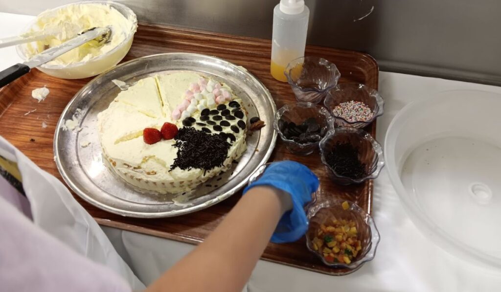 This image shows a cake decorating workshop on the Regal Princess cruise ship. A partially decorated cake sits on a tray, topped with various ingredients like marshmallows, strawberries, and sprinkles. Several small bowls of toppings, including sprinkles, chocolate chips, and nuts, are neatly arranged on the side, while a participant wearing a glove works on the decoration.