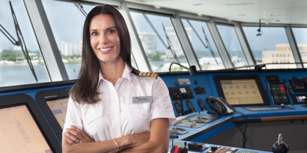 A confident female ship captain, identified by her epaulettes and name tag reading 'Captain', stands proudly in the ship's bridge with a backdrop of large windows showing a coastal view and advanced navigational equipment around her.